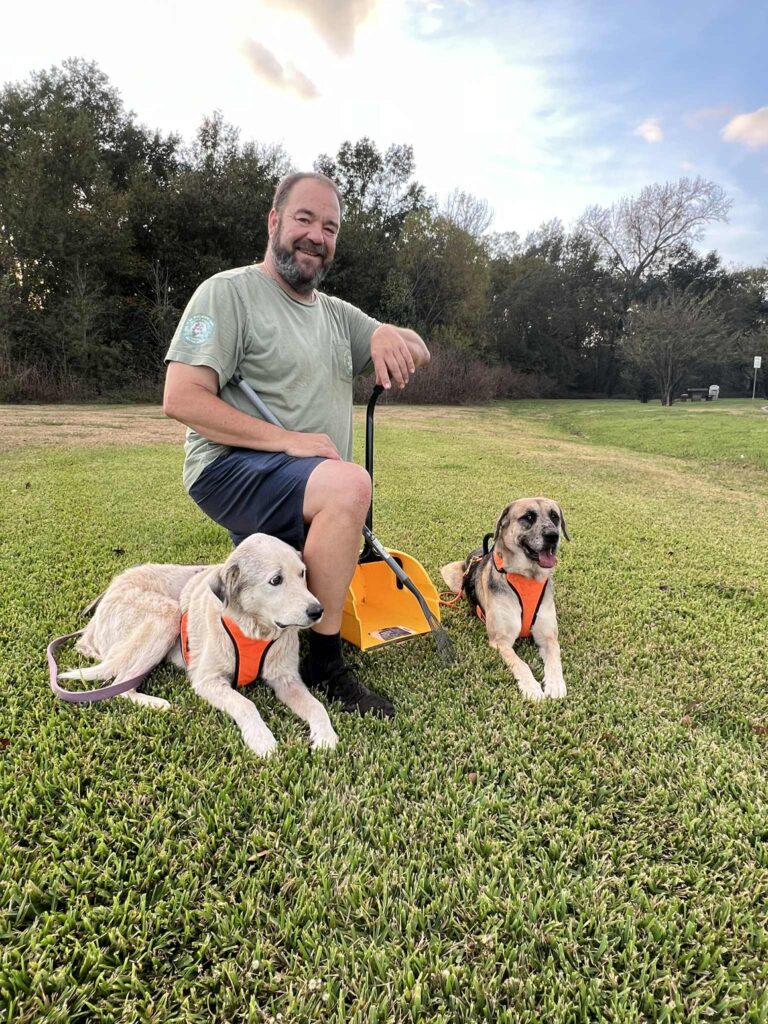 Duty Dog scoop technician sitting in a grassy yard with two service dogs wearing orange harnesses, ready for pet waste cleanup in East Texas.